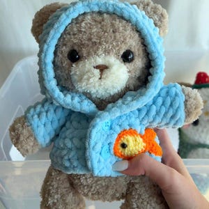 K&ouml;nnte beinhalten: Ein brauner Teddyb&auml;r mit wei&szlig;em Gesicht tr&auml;gt einen geh&auml;kelten blauen Kapuzenpullover mit einem gelben Fisch-Applikationen auf der Vorderseite.