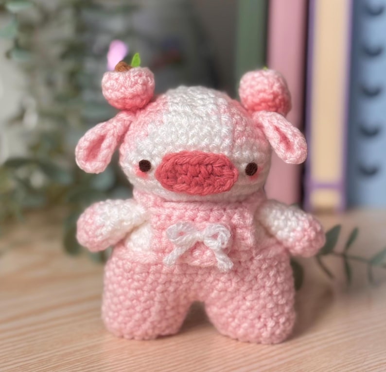 May include: A pink and white crocheted stuffed animal shaped like a cow with a pink nose and black eyes. The cow is wearing a pink and white crocheted outfit with a white bow.
