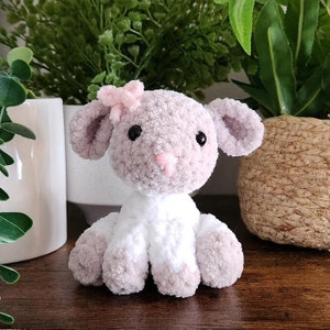 May include: A crocheted plush lamb toy with a pink bow, white fur, and a light purple face. The lamb is sitting on a wooden surface.