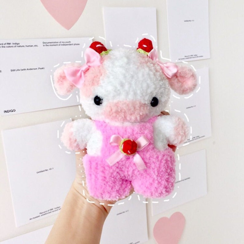 May include: A pink and white crocheted cow plush toy wearing a pink overall with a red bow and a strawberry on the front. The toy has black eyes and a pink nose.