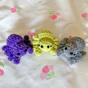 May include: Three handmade crochet spiders in purple, yellow-green, and gray. Each spider has eight legs and black safety eyes. The spiders are displayed on a white fabric with a floral pattern.