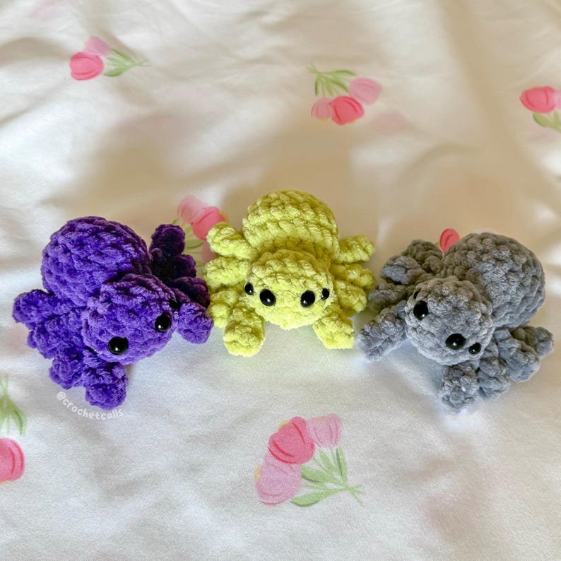 May include: Three handmade crochet spiders in purple, yellow-green, and gray. Each spider has eight legs and black safety eyes. The spiders are displayed on a white fabric with a floral pattern.