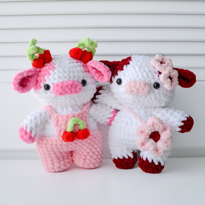 May include: Two crocheted cow plushies, one pink and white with a cherry design and the other red and white with a flower design.