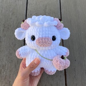 May include: A white crocheted plush toy shaped like a cow with pink ears, a pink nose, and black eyes. The toy is holding a pink crocheted flower in its right hand.