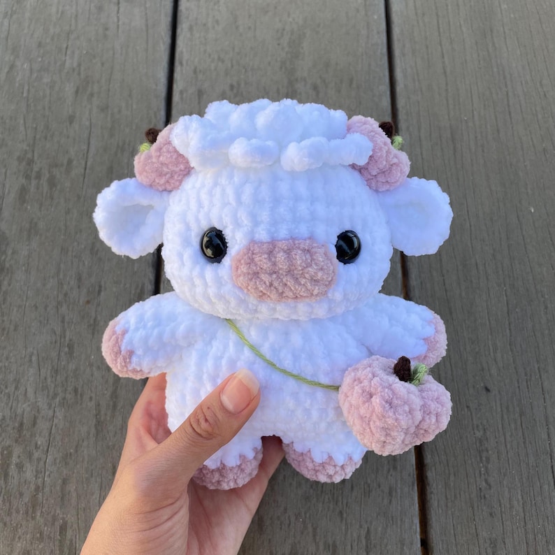 May include: A white crocheted plush toy shaped like a cow with pink ears, a pink nose, and black eyes. The toy is holding a pink crocheted flower in its right hand.