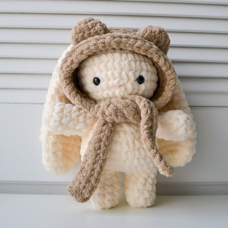 May include: A crocheted plush toy bunny wearing a brown hooded scarf. The bunny is cream coloured and has black eyes and a pink nose.