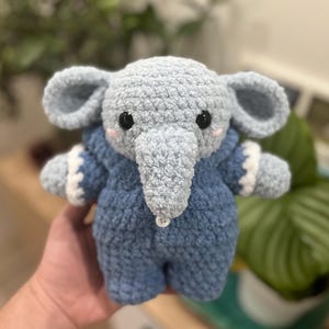 May include: A crocheted blue elephant plush toy wearing a blue and white crocheted onesie. The elephant has a small button on its chest.