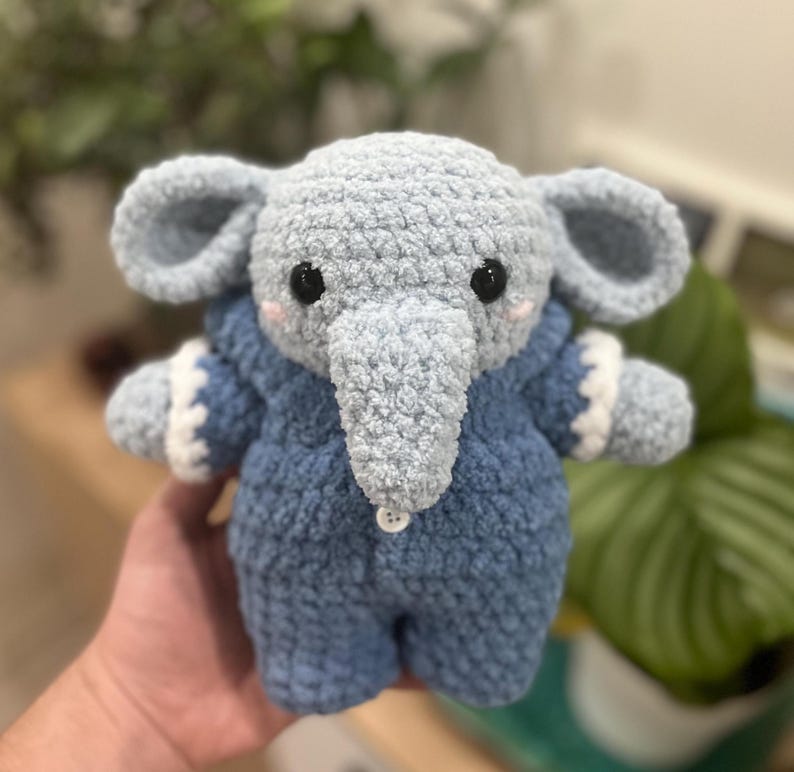 May include: A crocheted blue elephant plush toy wearing a blue and white crocheted onesie. The elephant has a small button on its chest.