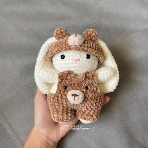 May include: A white crocheted bunny with brown crocheted ears and a brown crocheted hat. The bunny is holding a brown crocheted bear with black eyes. The text "Ducky Crochet Corner" is visible in the bottom right corner of the image.