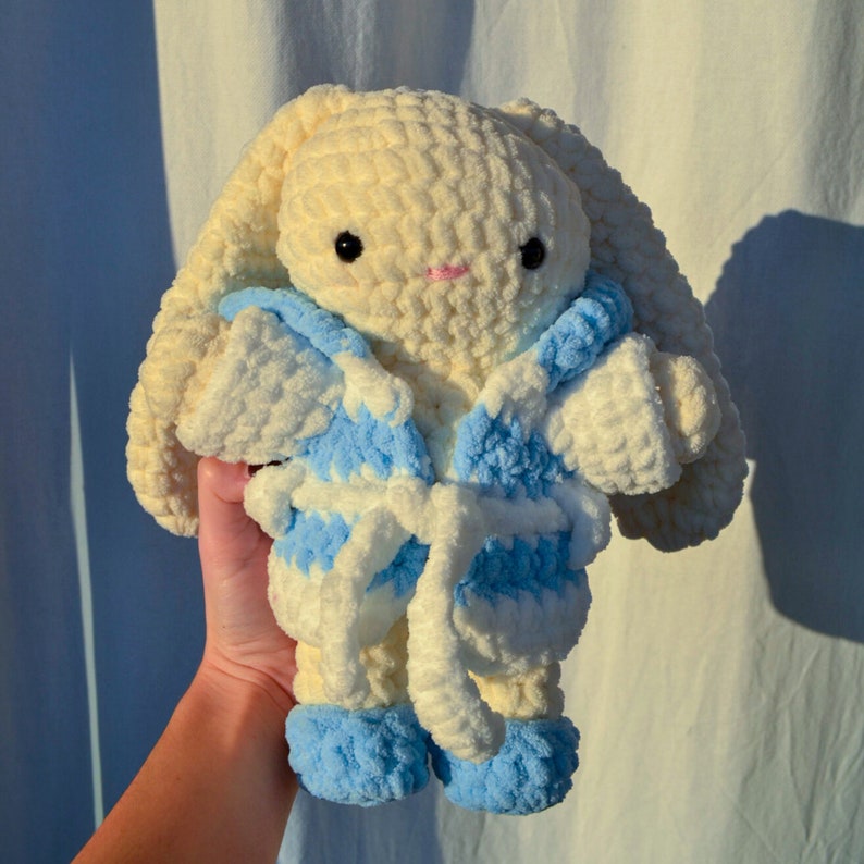 May include: A crocheted plush bunny wearing a blue and white robe. The bunny has a cream coloured body and black eyes.