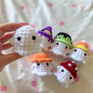 May include: A collection of handmade crochet ghosts, each with a unique hat. The ghosts are white with black eyes and pink cheeks. Hats are in various colors, including purple, orange, green, and red with white polka dots. The ghosts are small and adorable.