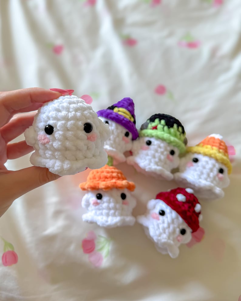 May include: A collection of handmade crochet ghosts, each with a unique hat. The ghosts are white with black eyes and pink cheeks. Hats are in various colors, including purple, orange, green, and red with white polka dots. The ghosts are small and adorable.