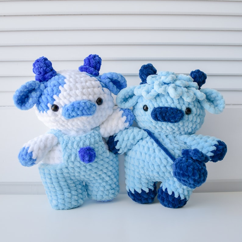 May include: Two crocheted stuffed animals shaped like cows. One is white and blue, the other is blue. Both have blue eyes and noses.