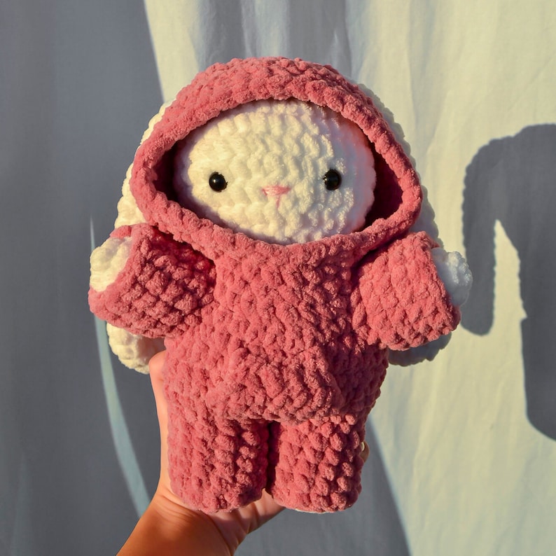 May include: A pink crocheted bunny wearing a pink hooded sweater. The bunny has white fur and black eyes.