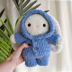 Pode incluir: Um coelho de croch&ecirc; branco vestindo um su&eacute;ter azul com capuz e bot&otilde;es. O coelho tem olhos pretos e um nariz rosa.