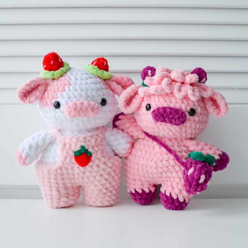 May include: Two crocheted stuffed toys shaped like cows. One is pink with a purple strawberry and the other is pink and white with a green strawberry.