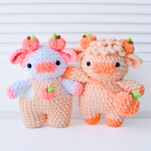 May include: Two crocheted plush toys shaped like cows with peach-colored fur and white accents. The cows have small black eyes and peach-colored noses. One cow is wearing a peach-colored overall with a peach-colored flower on the front. Both cows have peach-colored flowers on their heads.