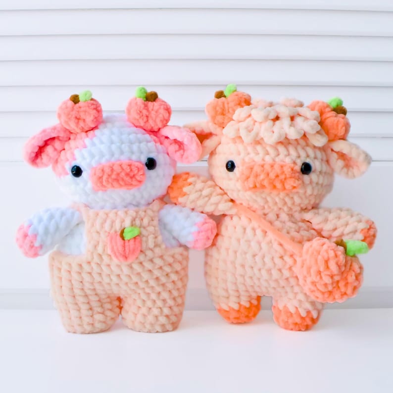 May include: Two crocheted plush toys shaped like cows with peach-colored fur and white accents. The cows have small black eyes and peach-colored noses. One cow is wearing a peach-colored overall with a peach-colored flower on the front. Both cows have peach-colored flowers on their heads.
