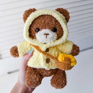 K&ouml;nnte beinhalten: Ein brauner Teddyb&auml;r, der eine gelbe geh&auml;kelte Jacke mit Kapuze, einem braunen Riemen und einer gelben geh&auml;kelten Tasche tr&auml;gt.