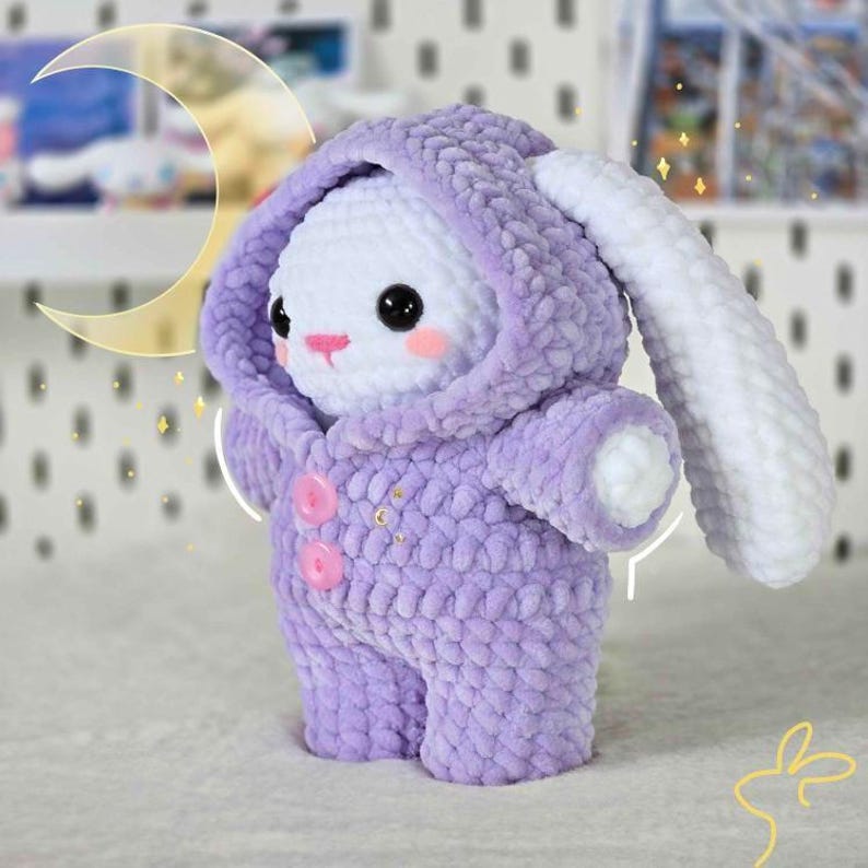 Pode incluir: Um coelho de pel&uacute;cia de croch&ecirc; vestindo um macac&atilde;o com capuz lavanda com dois bot&otilde;es rosa. O coelho tem pelo branco e uma cauda longa e branca.
