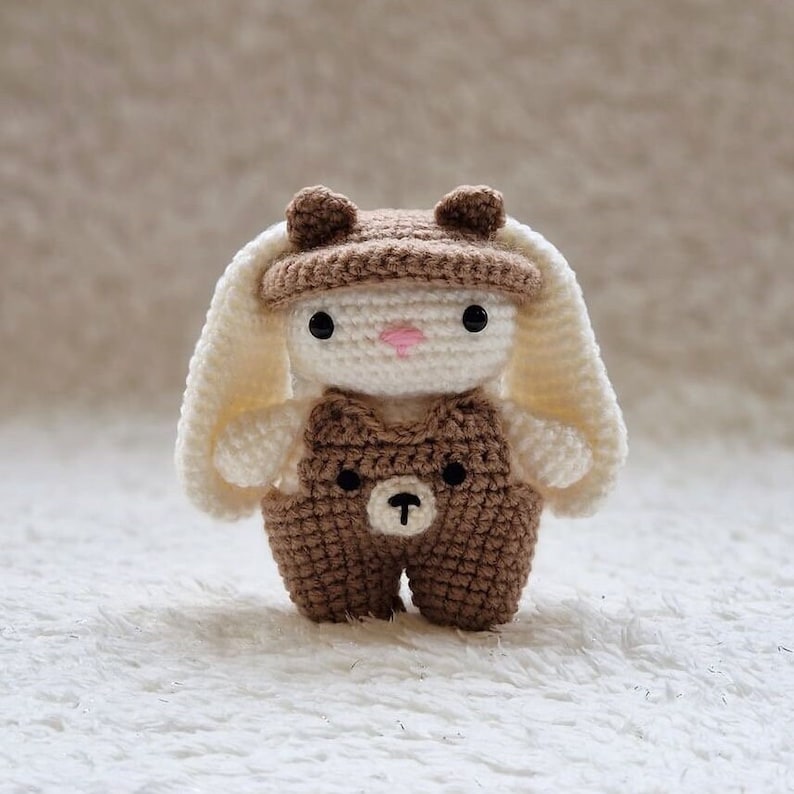 May include: A crocheted bunny toy wearing a brown hat and overalls with a bear face on the front. The bunny has white fur and pink nose.