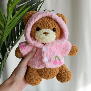 K&ouml;nnte beinhalten: Ein brauner geh&auml;kelter Teddyb&auml;r, der eine rosa geh&auml;kelte Kapuzenjacke mit gestickten Kirschen tr&auml;gt.