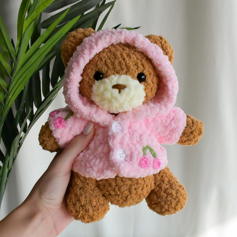 K&ouml;nnte beinhalten: Ein brauner geh&auml;kelter Teddyb&auml;r, der eine rosa geh&auml;kelte Kapuzenjacke mit gestickten Kirschen tr&auml;gt.