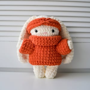 May include: A crocheted bunny toy wearing a red sweater and headband. The bunny has white fur and black eyes.