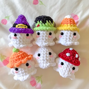 May include: Six handmade crochet ghosts with various hats. The hats include a purple witch hat, a green and black hat, a candy corn hat, an orange mushroom hat, and a red mushroom hat. Each ghost has black eyes and pink cheeks.