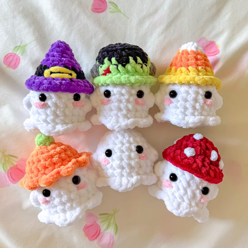 May include: Six handmade crochet ghosts with various hats. The hats include a purple witch hat, a green and black hat, a candy corn hat, an orange mushroom hat, and a red mushroom hat. Each ghost has black eyes and pink cheeks.