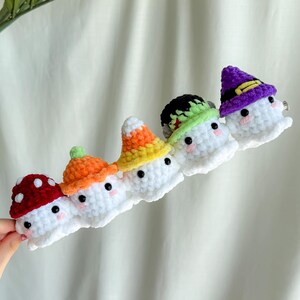 May include: A row of five white crochet ghost hair clips, each with a different colored hat. The hats are red with white spots, orange, yellow, black and green, and purple with a gold band. Each ghost has black eyes and pink cheeks.