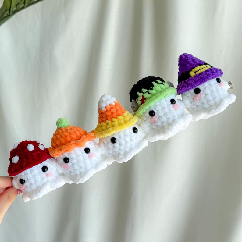 May include: A row of five white crochet ghost hair clips, each with a different colored hat. The hats are red with white spots, orange, yellow, black and green, and purple with a gold band. Each ghost has black eyes and pink cheeks.