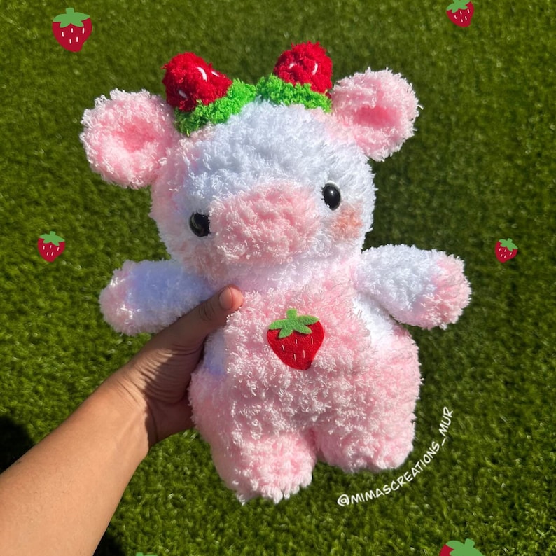 May include: A pink and white plush cow toy with a strawberry headband and a strawberry patch on its chest. The toy is being held by a hand.
