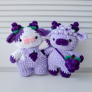 May include: Two crocheted plush toys shaped like cows with purple and white fur. The cow on the left has a white face and is wearing purple overalls with a grape design. The cow on the right is all purple and is holding a bunch of purple grapes.