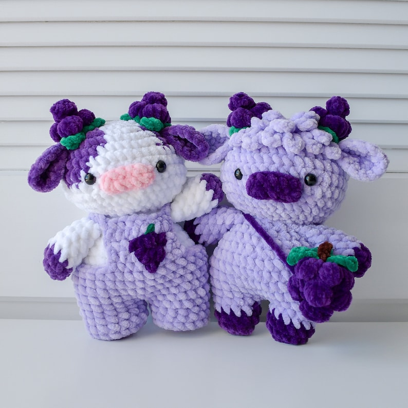 May include: Two crocheted plush toys shaped like cows with purple and white fur. The cow on the left has a white face and is wearing purple overalls with a grape design. The cow on the right is all purple and is holding a bunch of purple grapes.