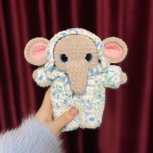 May include: A crocheted elephant wearing a blue and white hooded jacket with green accents. The elephant has a gray body, pink ears, and blue eyes.