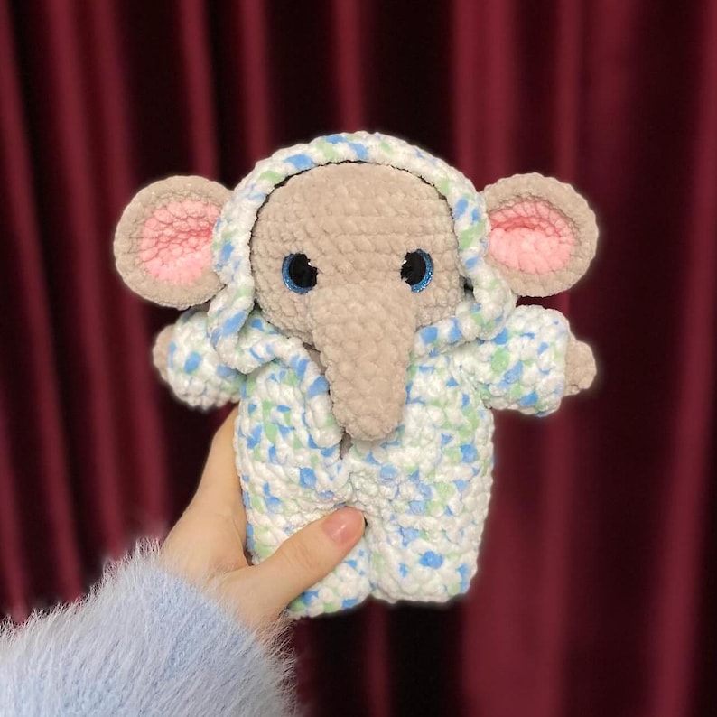 May include: A crocheted elephant wearing a blue and white hooded jacket with green accents. The elephant has a gray body, pink ears, and blue eyes.