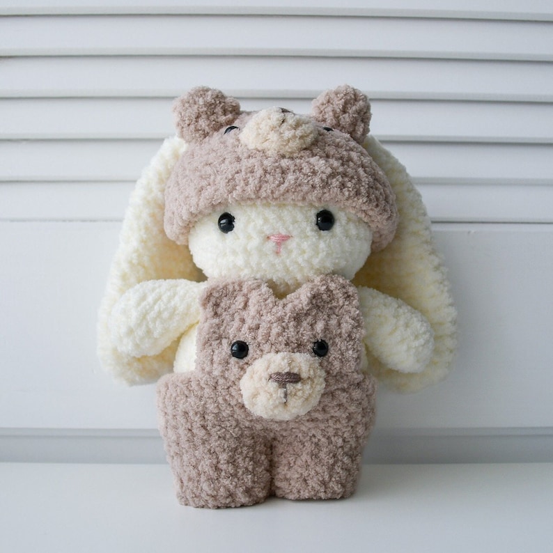 May include: A white crocheted bunny wearing a brown crocheted bear hat and a brown crocheted bear outfit.