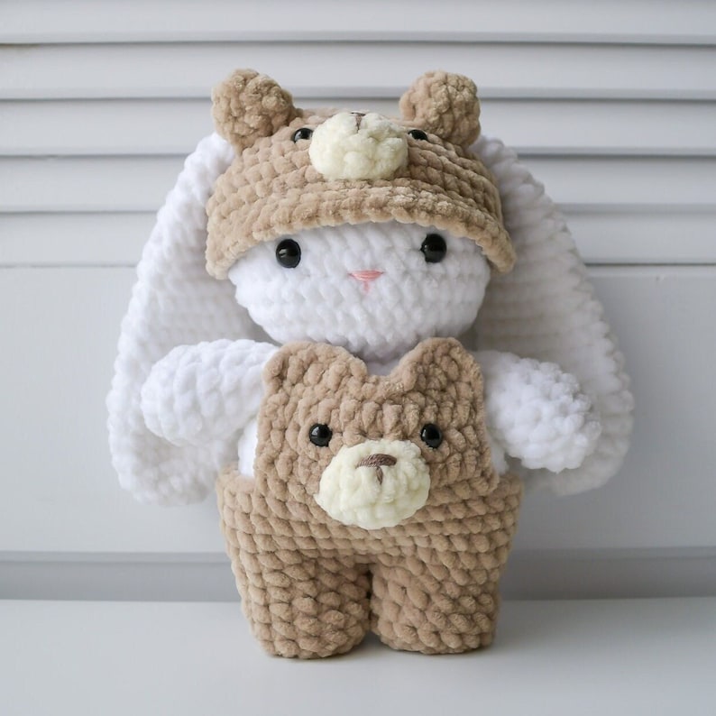 May include: A white crocheted bunny wearing a brown crocheted bear hat and a brown crocheted bear outfit. The bunny has black eyes and a pink nose.