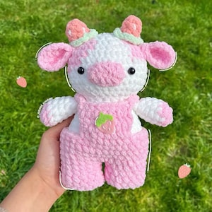 May include: A crocheted plush toy cow in pink and white with a strawberry design on its overalls. The cow has black eyes and a pink nose.