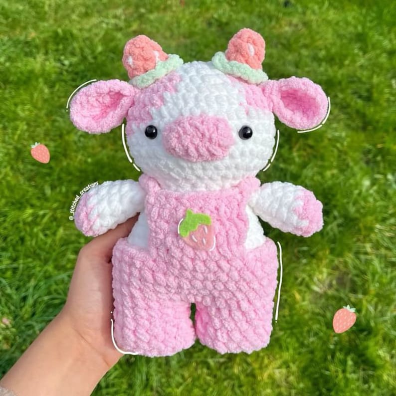 May include: A crocheted plush toy cow in pink and white with a strawberry design on its overalls. The cow has black eyes and a pink nose.
