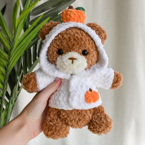 K&ouml;nnte beinhalten: Ein brauner geh&auml;kelter Teddyb&auml;r, der eine wei&szlig;e geh&auml;kelte Kapuzenjacke mit einem kleinen orangefarbenen geh&auml;kelten K&uuml;rbis auf der Vorderseite tr&auml;gt. Der B&auml;r tr&auml;gt einen kleinen orangefarbenen geh&auml;kelten K&uuml;rbis auf dem Kopf.