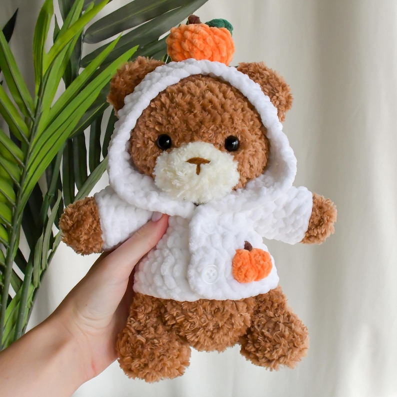 K&ouml;nnte beinhalten: Ein brauner geh&auml;kelter Teddyb&auml;r, der eine wei&szlig;e geh&auml;kelte Kapuzenjacke mit einem kleinen orangefarbenen geh&auml;kelten K&uuml;rbis auf der Vorderseite tr&auml;gt. Der B&auml;r tr&auml;gt einen kleinen orangefarbenen geh&auml;kelten K&uuml;rbis auf dem Kopf.