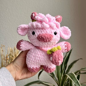 May include: A pink crocheted stuffed animal shaped like a cow with a pink strawberry in its hand. The cow has a pink nose and black eyes.