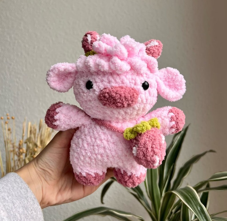 May include: A pink crocheted stuffed animal shaped like a cow with a pink strawberry in its hand. The cow has a pink nose and black eyes.