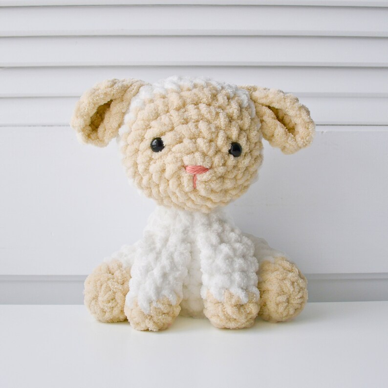May include: A crocheted plush toy lamb with a white body and beige head and legs. The lamb has black eyes and a pink nose.