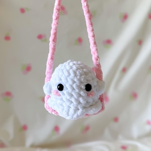 May include: A handmade white crochet ghost-shaped bag with black eyes and pink cheeks. The bag has a pink strap and trim, creating a soft, plush appearance. The background is a light pink and white floral pattern.