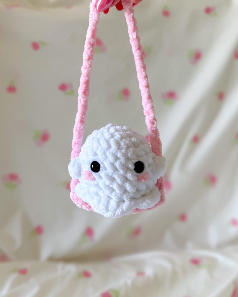 May include: A handmade white crochet ghost-shaped bag with black eyes and pink cheeks. The bag has a pink strap and trim, creating a soft, plush appearance. The background is a light pink and white floral pattern.
