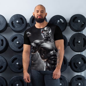 Może przedstawiać: Czarny t-shirt z nadrukiem graficznym przedstawiającym robota z czerwonym okiem. Postać jest muskularna i ma szczegółowy projekt.
