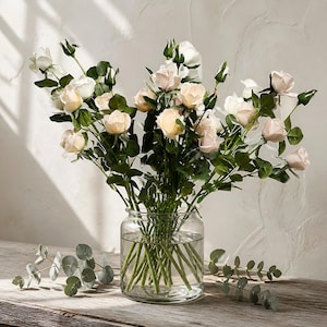May include: A clear glass vase filled with a bouquet of artificial roses in shades of cream, pale pink, and white. The roses feature green leaves and stems, with several small buds. Two sprigs of eucalyptus foliage are placed at the base of the vase.